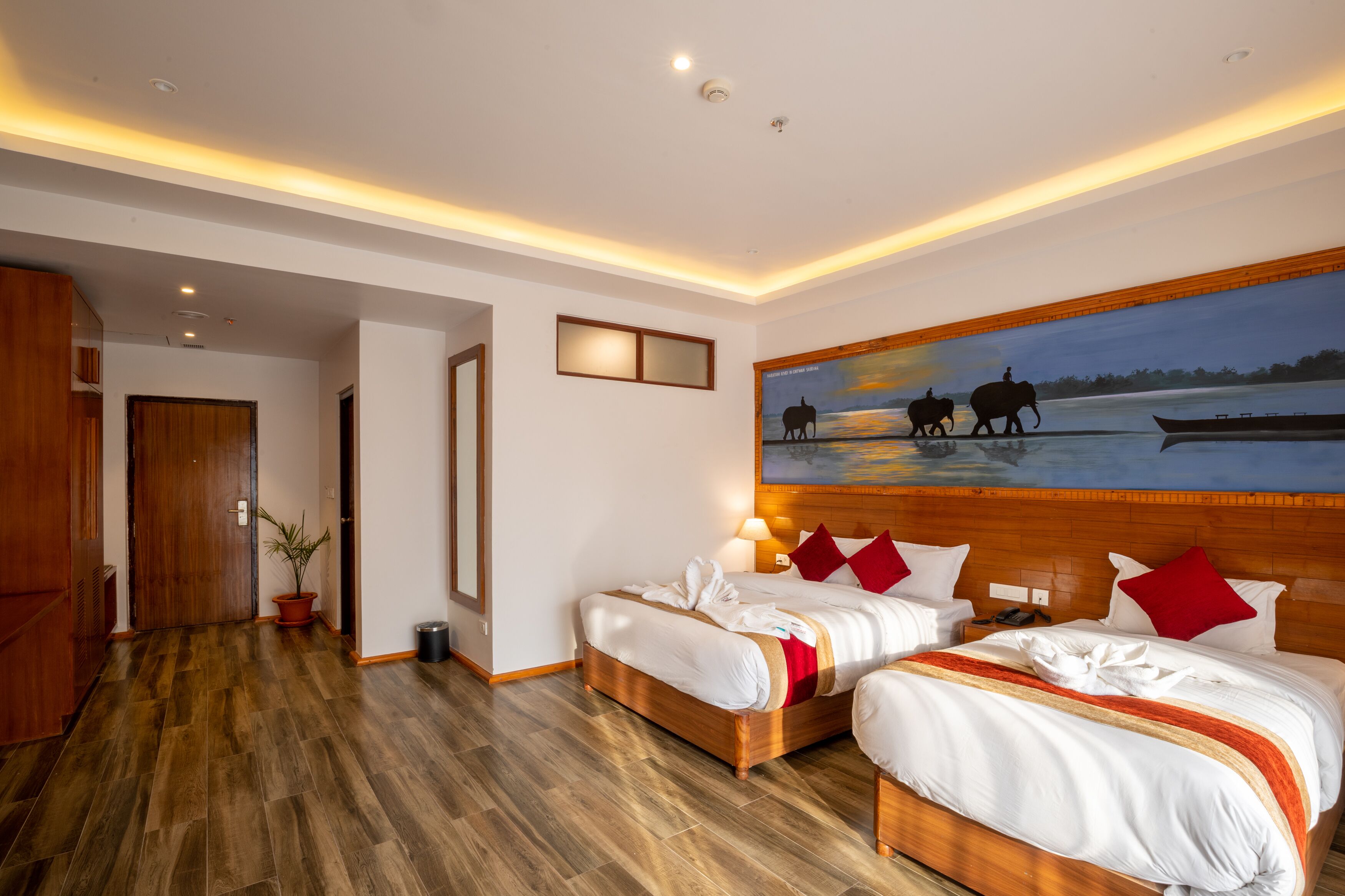 A hotel room with two beds, wooden flooring, a painting of elephants, and a wooden headboard.
