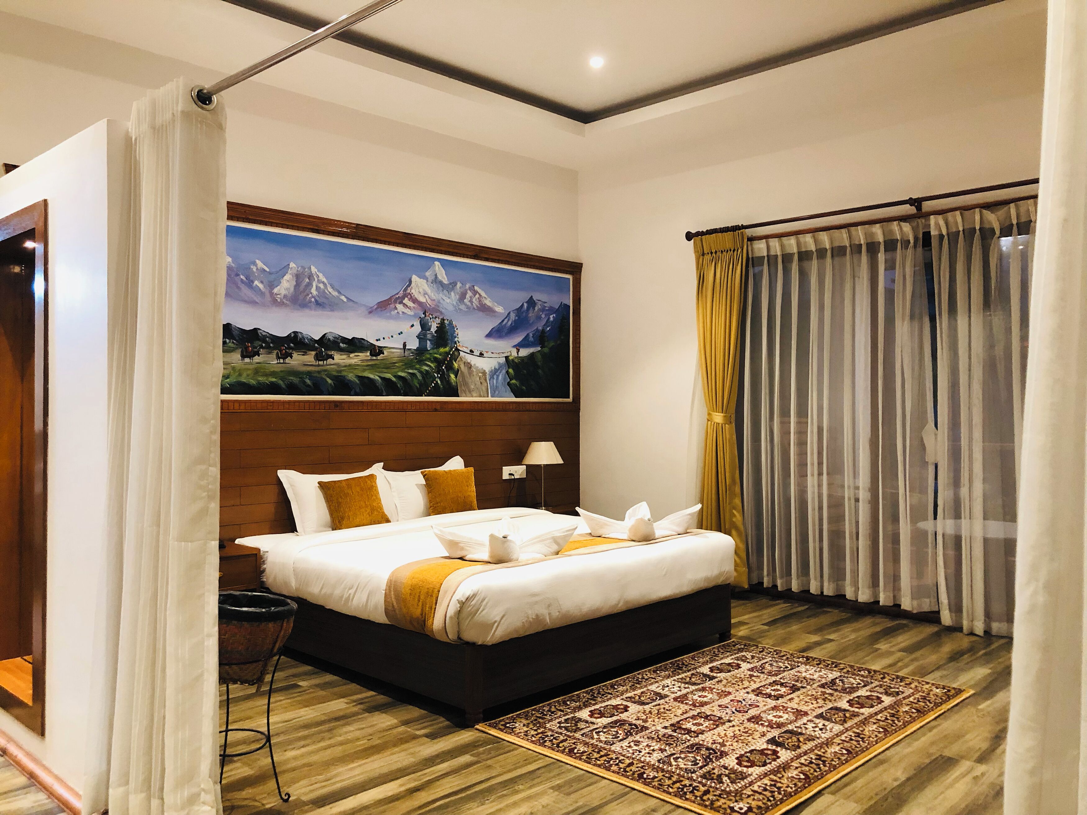 A hotel room with a large bed, a wooden headboard, a painting of mountains, a window with curtains, and a patterned rug.