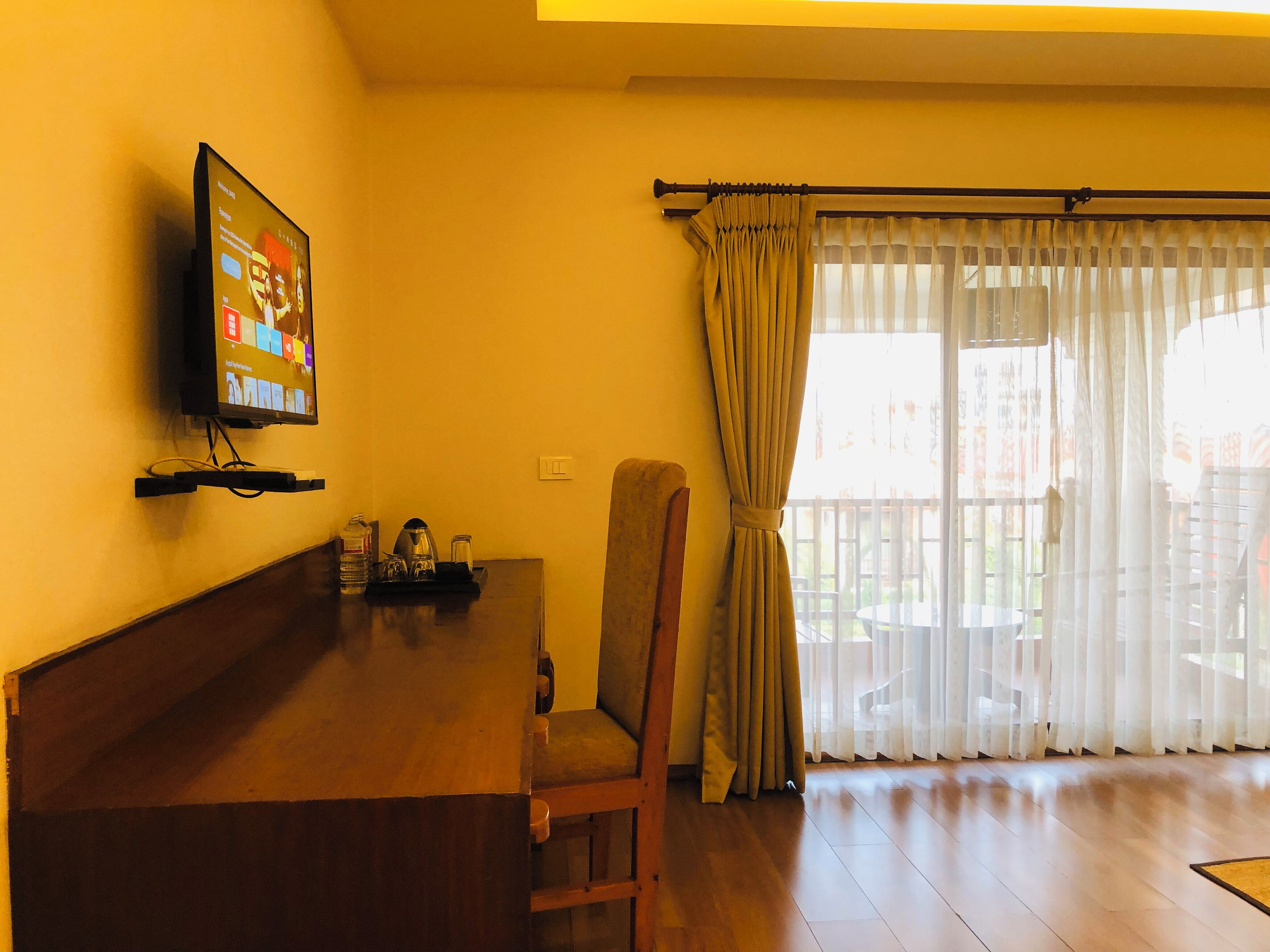 A living room with a wooden coffee table, a television mounted on the wall, a chair, and a window with curtains.