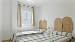 Un dormitorio con dos camas, una ventana con cortinas, una mesita de noche con lámpara y un cabecero tejido fijado a la pared.