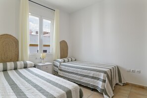 A bedroom with two beds, a window with curtains, a bedside lamp, and a wall socket.