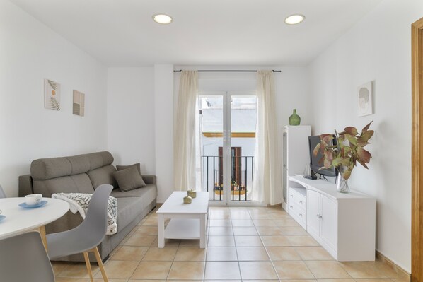 A modern living room with a sofa, a dining table, a TV on a cabinet, and a balcony with curtains.