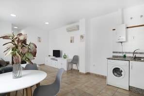 A modern kitchen with a washing machine, a dining area with a table and chairs, and a living area with a TV and a plant.