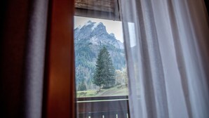 Ein Fenster mit Blick auf eine Berglandschaft, Vorhänge sind teilweise zurückgezogen, und eine Holzbalkendecke.