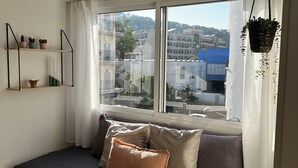 A cozy bedroom with a wooden bed, a window with a view of buildings, and a shelf with plants. - Charming apartment in Tossa de Mar (Tossa de Mar)
