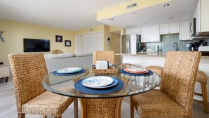 Una cocina moderna con armarios blancos, un refrigerador de acero inoxidable y un comedor con sillas de mimbre y una mesa de vidrio.