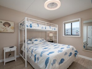 Quarto com beliche, colcha branca e azul, mesa de cabeceira, prateleira na parede, janela com vista e luminária no teto.