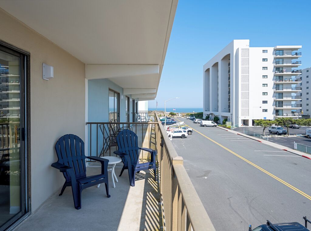 Ein Balkon mit zwei Stühlen und einem Tisch, der auf eine Straße mit Autos und einem Gebäude im Hintergrund blickt.
