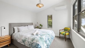 Un dormitorio con cama, mesita de noche, silla, ventana y aire acondicionado.