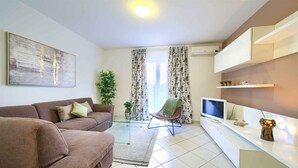 Ein Wohnzimmer mit einer Couch, einem Sessel, einem Fernseher, einem Glastisch und einer Pflanze.