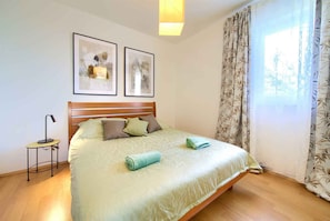 A neatly made bed with a patterned bedspread, two framed artworks above it, a bedside lamp, and a window with curtains.