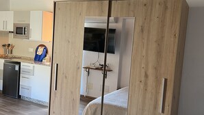 A bedroom with a bed, wooden wardrobe, and a kitchenette in the background.