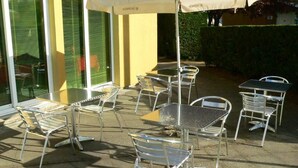 Mobiliario de patio exterior con sillas y mesas de metal, una sombrilla cubierta y un edificio con contraventanas verdes al fondo.