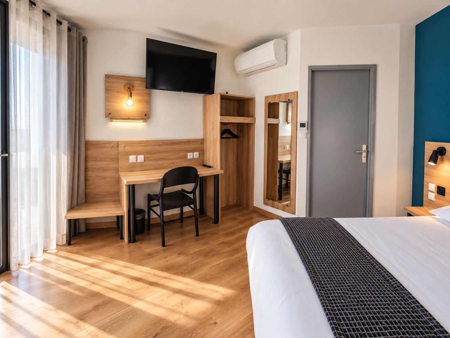 Una habitación de hotel con cama, escritorio con silla, televisor montado en la pared, unidad de aire acondicionado y puerta con espejo.