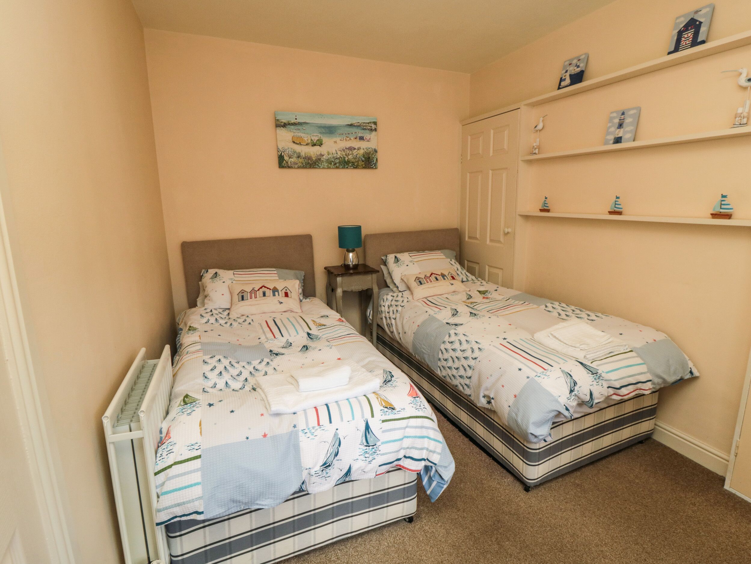 A bedroom with two single beds, a nightstand with a lamp, a shelf with decorative items, and a painting on the wall.