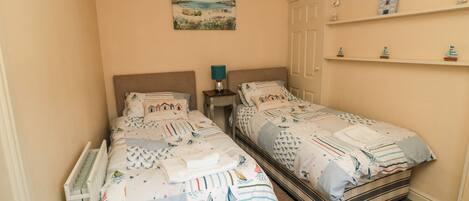 A bedroom with two single beds, a nightstand with a lamp, a shelf with decorative items, and a painting on the wall.