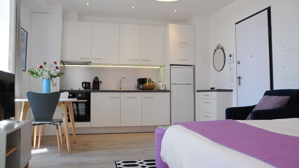 Een moderne, compacte hotelkamer met kitchenette, een bed met paars beddegoed en een zithoek met een blauwe stoel.