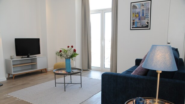 Una sala de estar moderna con un televisor de pantalla plana, un sofá azul, una mesa de centro con un jarrón de flores y una lámpara.