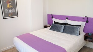 Una cama bien hecha con un cabecero morado, un cartel enmarcado en la pared y una mesita de noche con un libro.