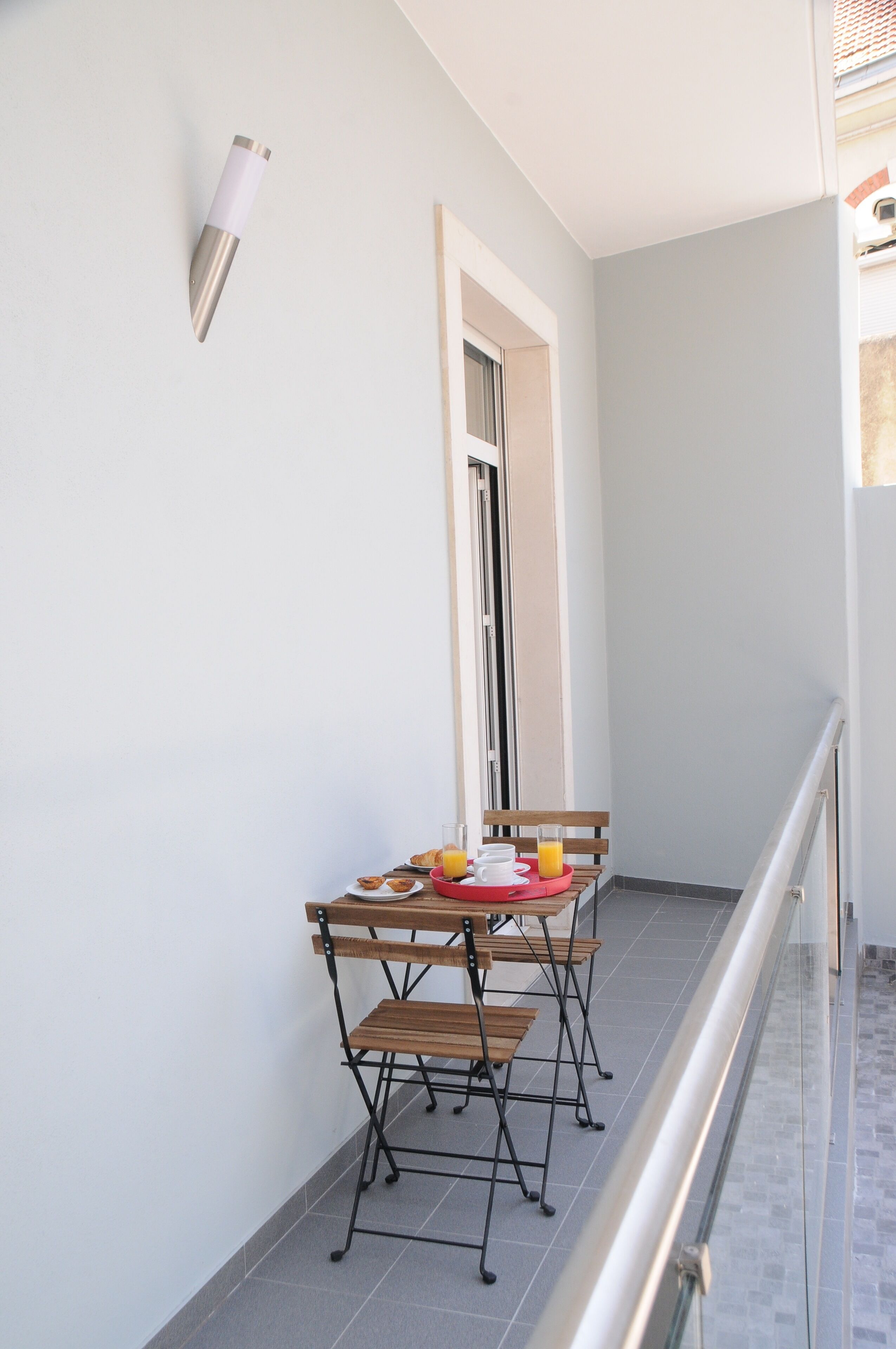 A balcony with a table and chairs, a small table with a tray of food and drinks, and a wall-mounted light fixture.
