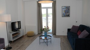 Una sala de estar moderna con un televisor de pantalla plana, una mesa de centro redonda con un florero, un sofá azul oscuro y un ventanal con cortinas.