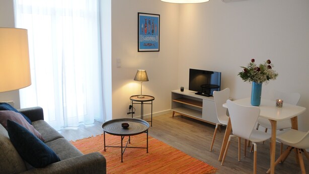 Ein modernes Wohnzimmer mit einer Couch, einem kleinen runden Tisch, einem Fernseher und einem Essbereich mit Stühlen und einer Blumenvase.