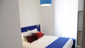 Ein ordentlich bezogenes Bett mit blauem Kopfteil, weißer Bettwäsche und einem roten Zierkissen.