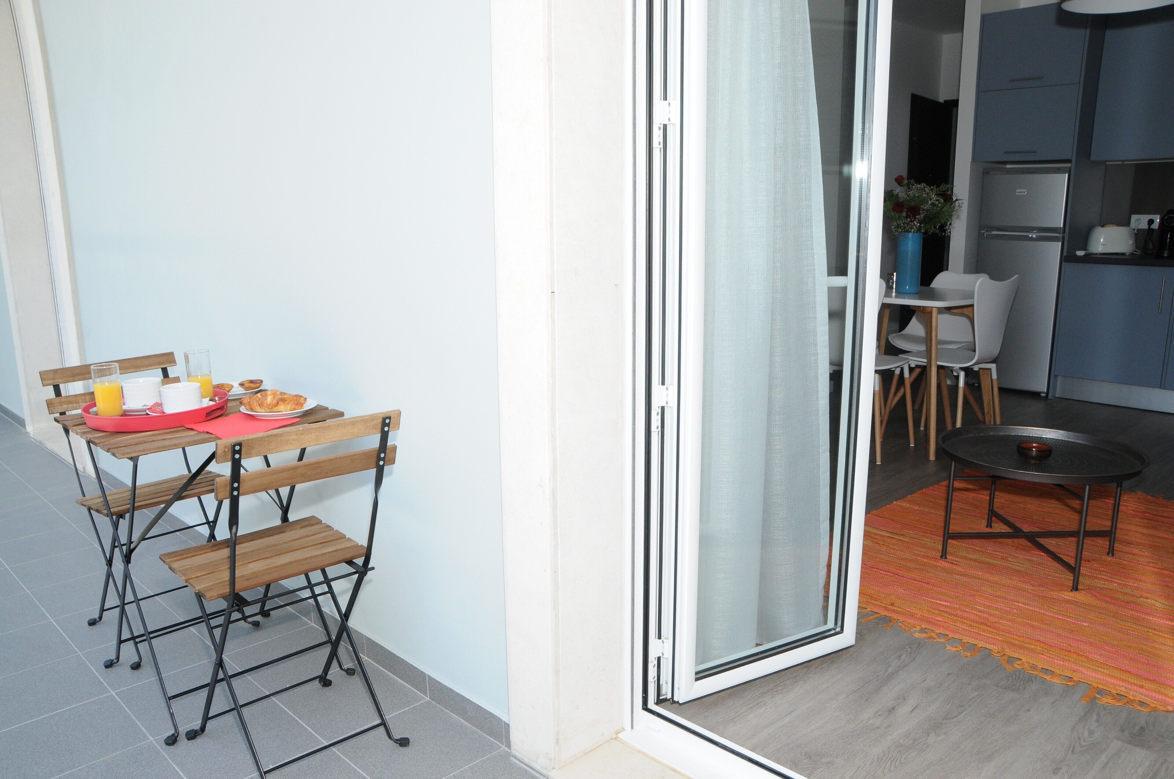 Un espacio habitable compacto con zona de comedor que incluye una mesa y sillas plegables, una pequeña cocina con refrigerador, y un vidrio corredizo que da a una habitación contigua.