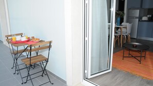 Ein kompakter Wohnbereich mit Essbereich, ausgestattet mit klappbarem Tisch und Stühlen, einer kleinen Küche inklusive Kühlschrank sowie einer Glastür, die in einen angrenzenden Raum führt.