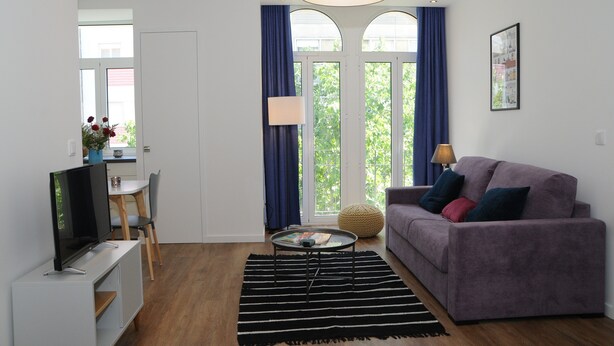 A modern living room with a flat-screen TV, a sofa, a coffee table, and a dining area with a chair and table. - Charming Comfortable Apartment with Balcony 1 (Lisboa)