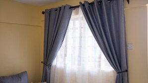 A room with a window covered by grey curtains, a white tiled wall, and a grey sofa with a striped pillow. - Letai Homes Furnished Apartment (Nairobi)