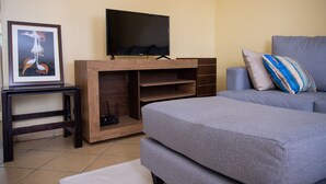 A modern living room with a flat-screen TV mounted on a wooden entertainment unit, a grey sofa with a striped pillow, and a framed artwork on the wall. - Letai Homes Furnished Apartment (Nairobi)
