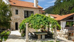 A stone house with a tiled roof, a covered patio with a table and benches, and a garden with various plants. - A Palleira: Antiguo Pajar Reformado al Lado del río (Pobra do Caramiñal)