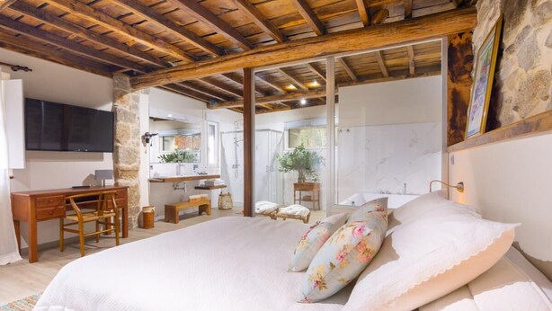 A bedroom with a large bed, wooden ceiling beams, a stone wall, a desk, and a bathroom visible through a glass door. - A Palleira: Antiguo Pajar Reformado al Lado del río (Pobra do Caramiñal)