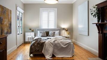 A bedroom with a bed, bedside lamps, a radiator, a window with shutters, and a plant on a stand.