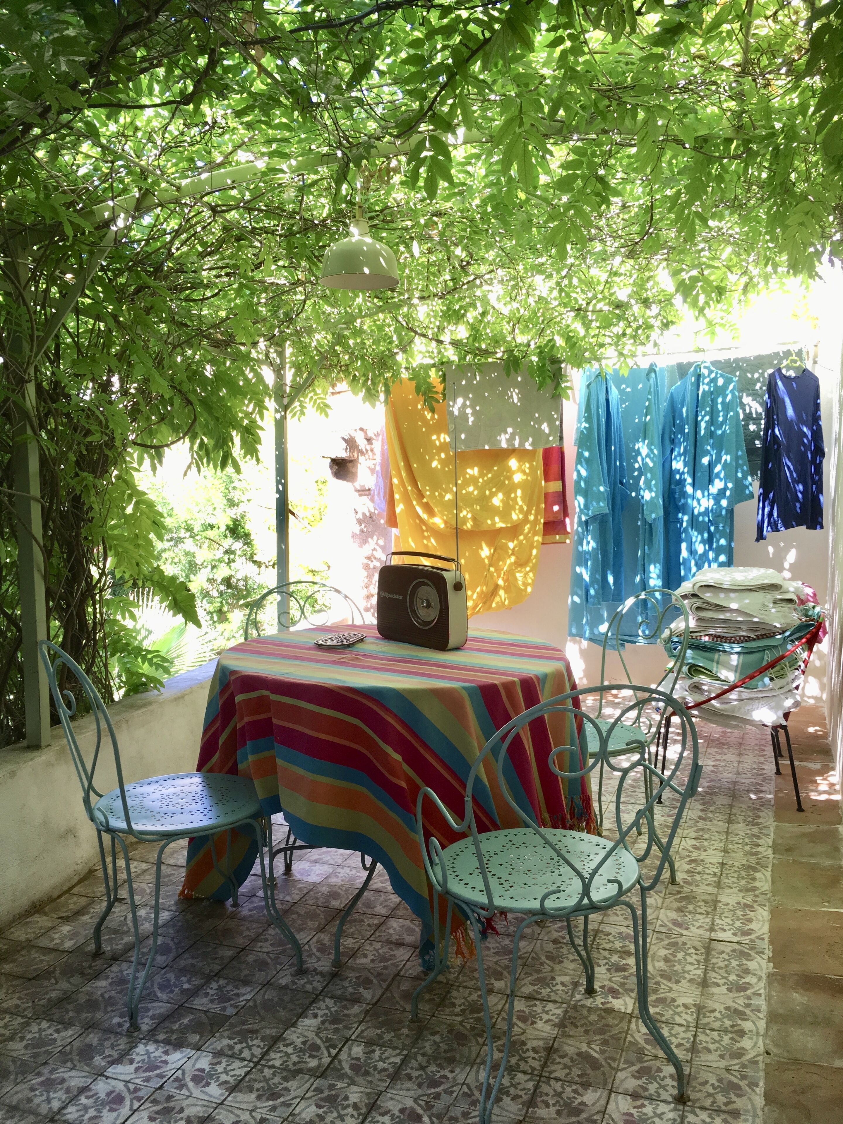 Une terrasse extérieure avec une table recouverte d’une nappe à rayures, des chaises en métal, une radio et des vêtements suspendus.