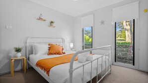 A bedroom with a white bed, a small bedside table, a nightstand, a window with a view of greenery, and a star decoration on the wall.
