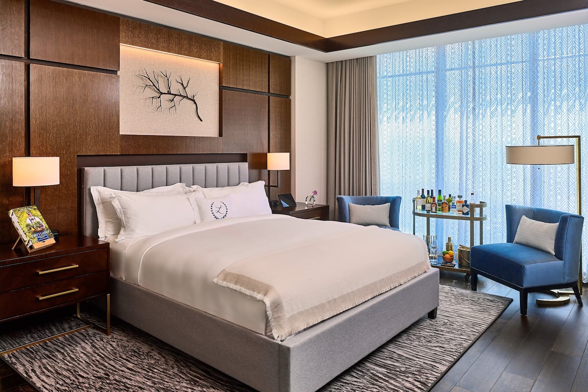 A hotel room with a large bed, a nightstand with a lamp, a chair, a small table with bottles, and a window with patterned curtains.