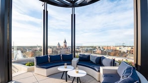A rooftop terrace with a curved seating area, two small round tables, and a view of a cityscape including a clock tower. - Laurel Hotel and Spa (Auburn)