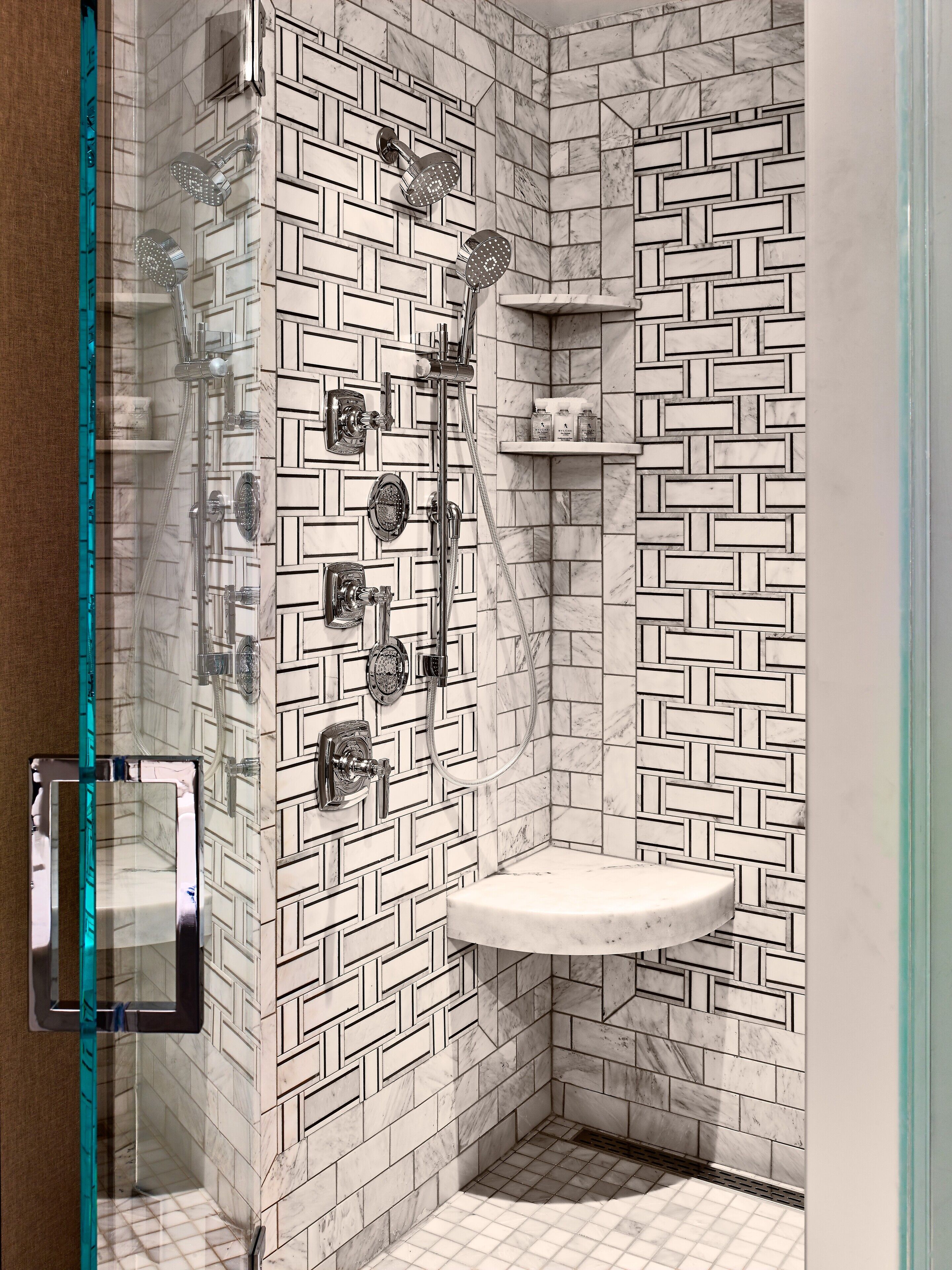 Un baño con pared de azulejos de diseño geométrico, cabina de ducha de vidrio, lavamanos de mármol y estante empotrado con artículos de aseo.