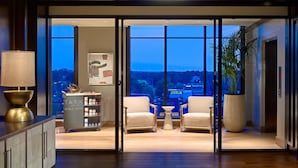 A modern hotel room with a large window offering a city view, a bar area with a minibar, and a seating area with two chairs and a small table. - Laurel Hotel and Spa (Auburn)