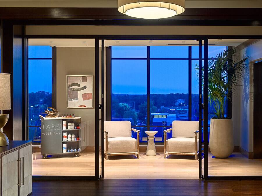 Habitación de hotel moderna con un ventanal amplio que ofrece vistas a la ciudad, una zona de bar con minibar, y una sala de estar con dos sillas y una mesita.
