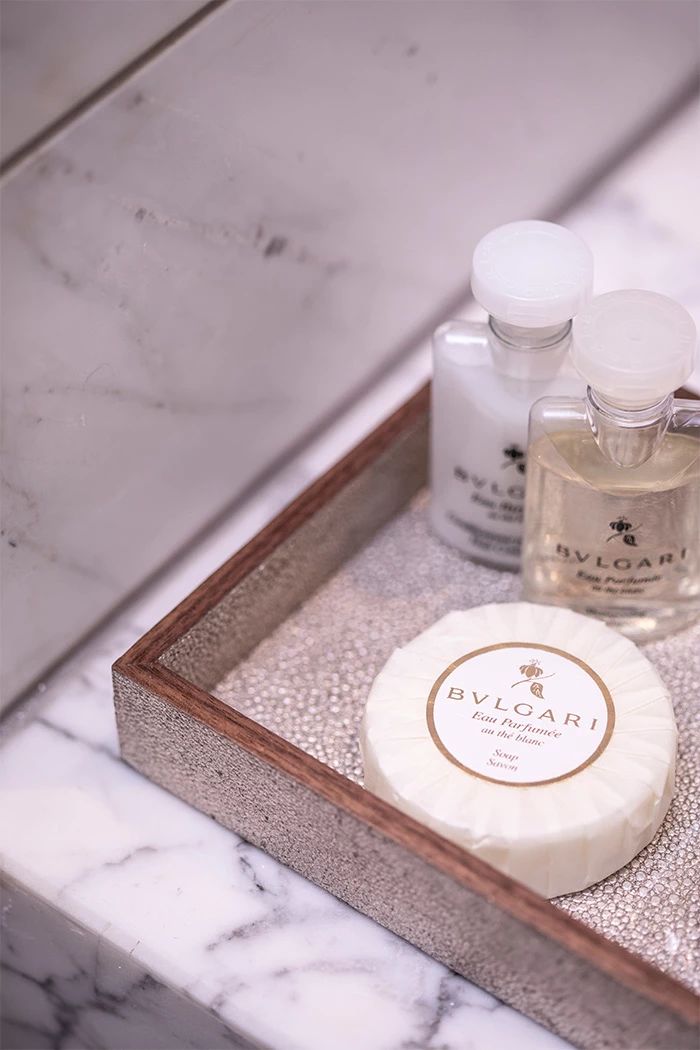 A wooden tray with a marble surface containing a round soap bar labeled "BVLOGARI" and two bottles of perfume.