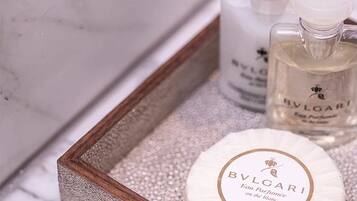 A wooden tray with a marble surface containing a round soap bar labeled "BVLOGARI" and two bottles of perfume.