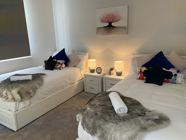 Two single beds with white bedding and colorful pillows, each with a rolled-up towel, are situated in this stylish room with a large window offering a view of the outside.