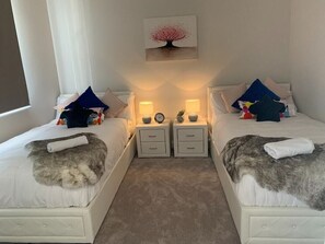 Two single beds with matching headboards, each with a white comforter and a throw blanket, are accompanied by bedside tables with lamps and clocks.