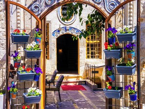 Área externa com assentos, cadeiras e um tapete vermelho, cercada por plantas em vasos e um arco decorativo.