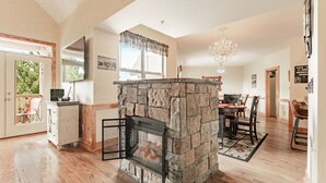 A spacious living room with a stone fireplace, wooden flooring, a dining area with a chandelier, and a view of the outdoors through large windows.