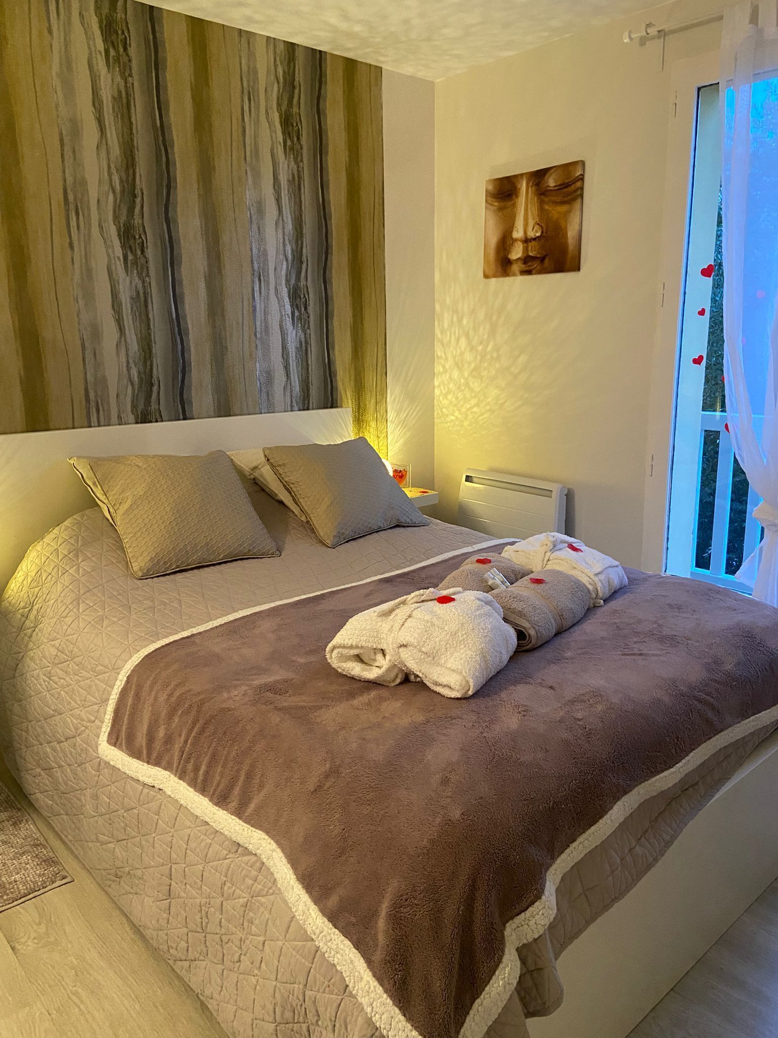 A bedroom with a large bed, a headboard with a wood-like texture, a bedside table with a lamp, and a window with curtains.