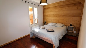 A bedroom with a wooden headboard, a bed with a patterned bedspread, a bedside table with a lamp, and a window with blinds.